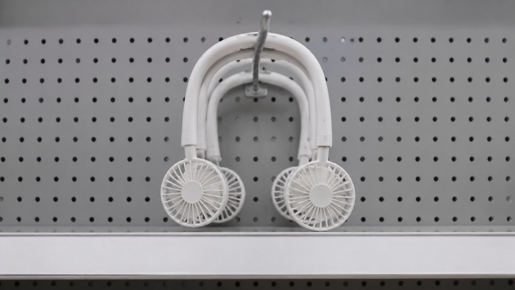 A white bladed neck fan hanging from a peg hook on a plain store pegboard (illustration)
